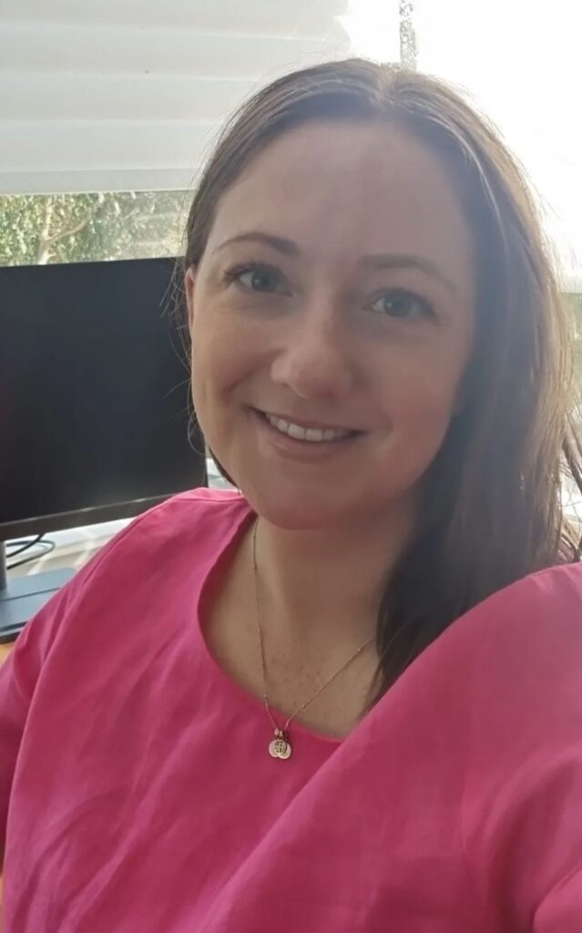 Stephanie Hazeltine wearing a pink top and smiling in front of her computer monitor