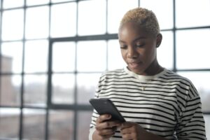 A woman standing in front of a window looks at her phone screen as though reading a text message.